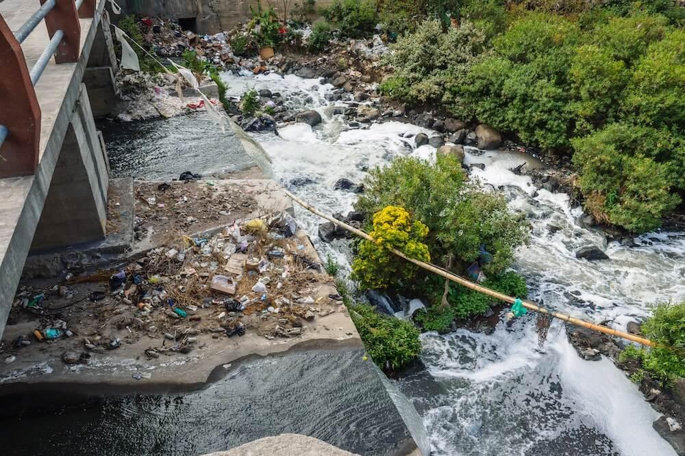 A bunch of plastic bottles and trash in a creek and plastic bags in the surrounding grass.